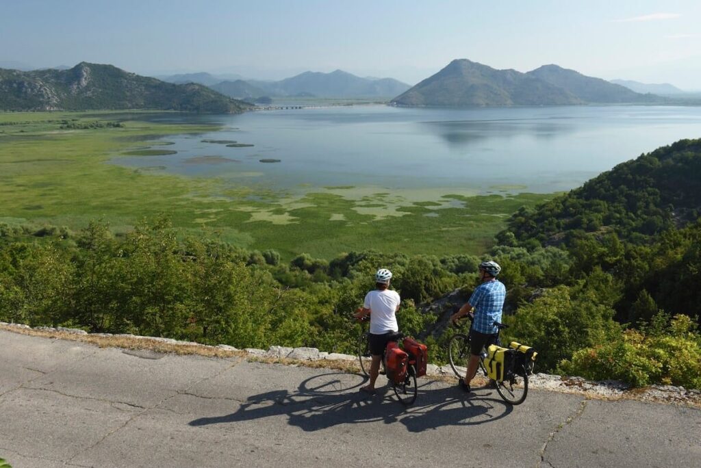 Two cyclists pause on a hilltop, admiring the expansive view of a lush green landscape and a tranquil lake surrounded by distant mountains.
